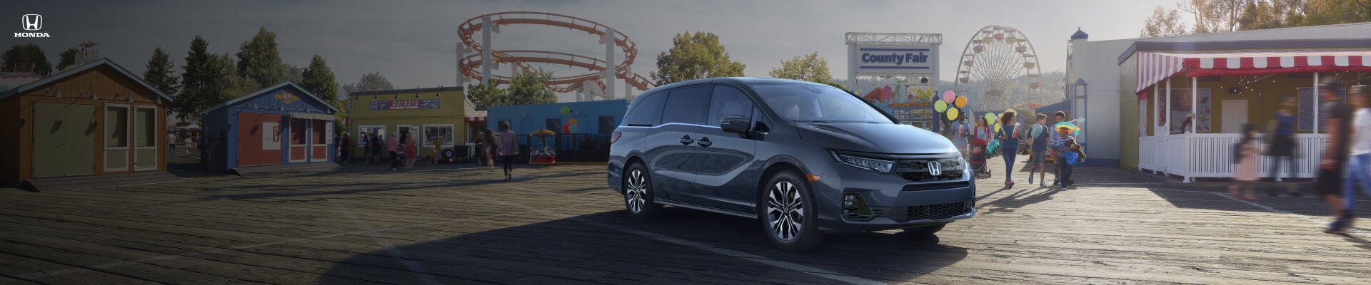 Front three-quarter view of a 2026 Honda Odyssey Elite in Smoke Blue Pearl parked near a county fairground with people.