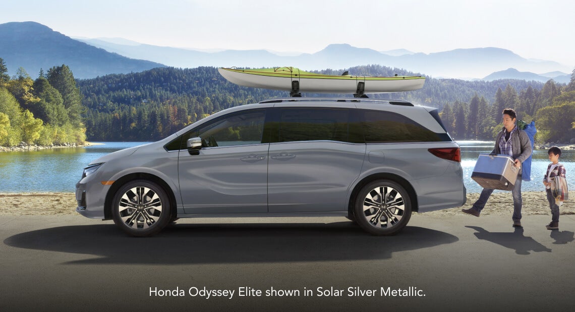 Side profile of a 2026 Honda Odyssey Elite in Solar Silver Metallic with kayak near a man and child by a lake.